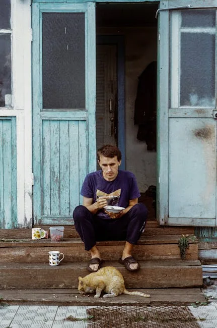 A man eating on wooden steps outside a rustic house with a teal door, a tabby cat sitting beside him.